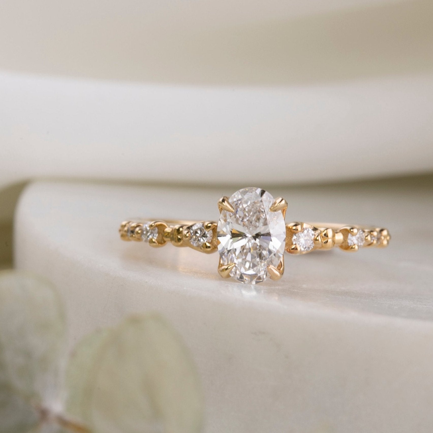 An oval-cut lab diamond ring with accent diamonds set in a yellow gold band, displayed on a white ceramic surface.