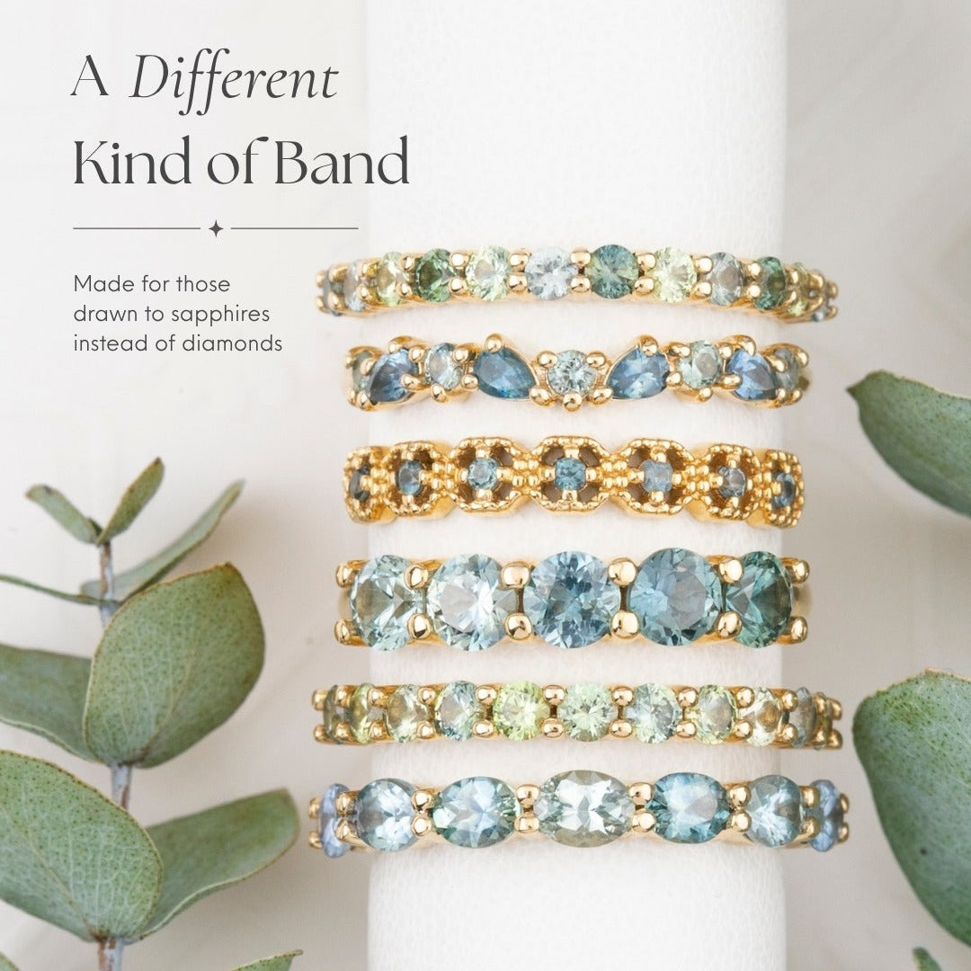 Five sapphire band rings in different colored stones and gold settings, displayed on a white cylindrical stand with eucalyptus leaves on both sides. Text on the left reads 'A Different Kind of Band' and 'Made for those drawn to sapphires instead of diamonds'.