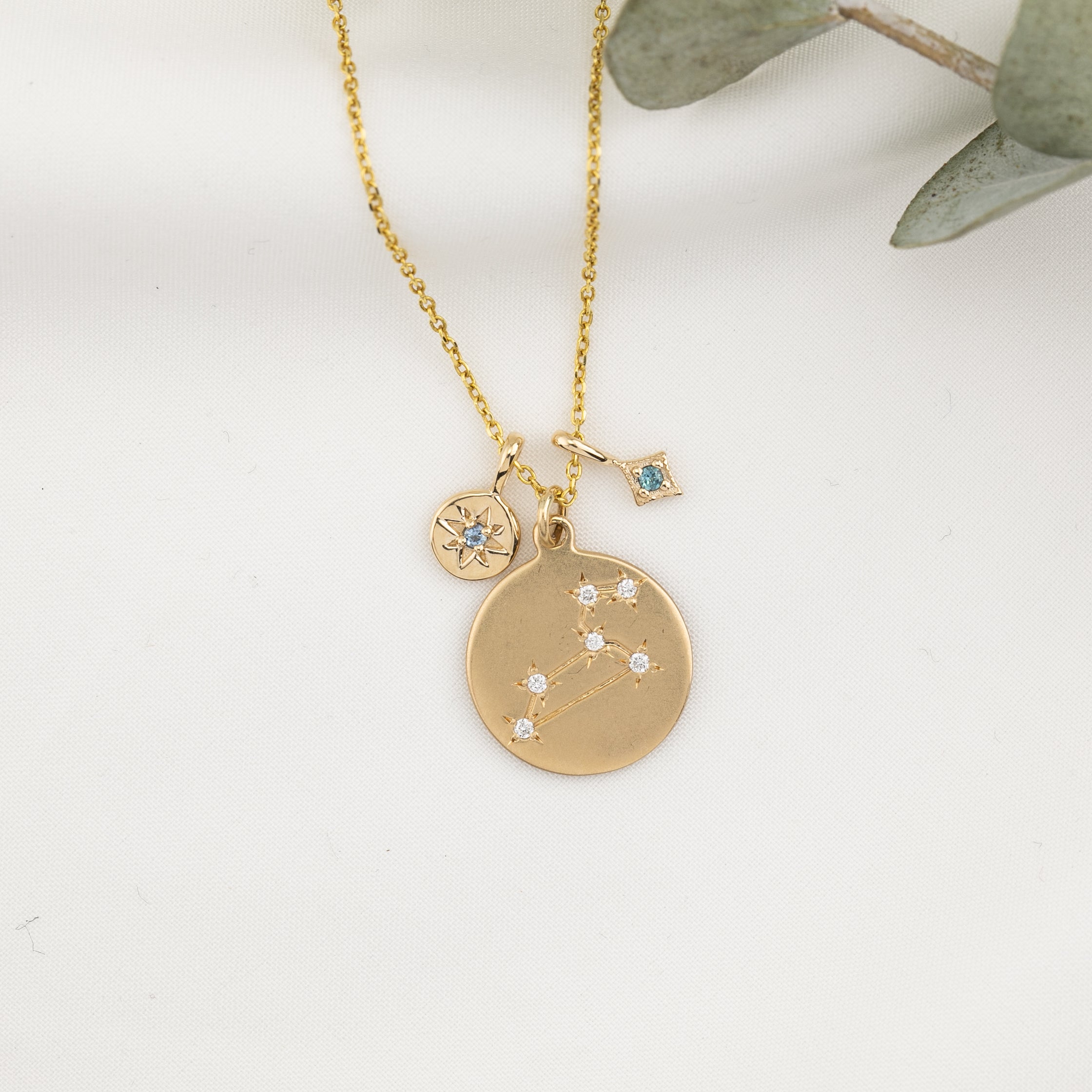 Gold necklace with constellation disc, initial star charm, and blue bezel-set birthstone charm on white background