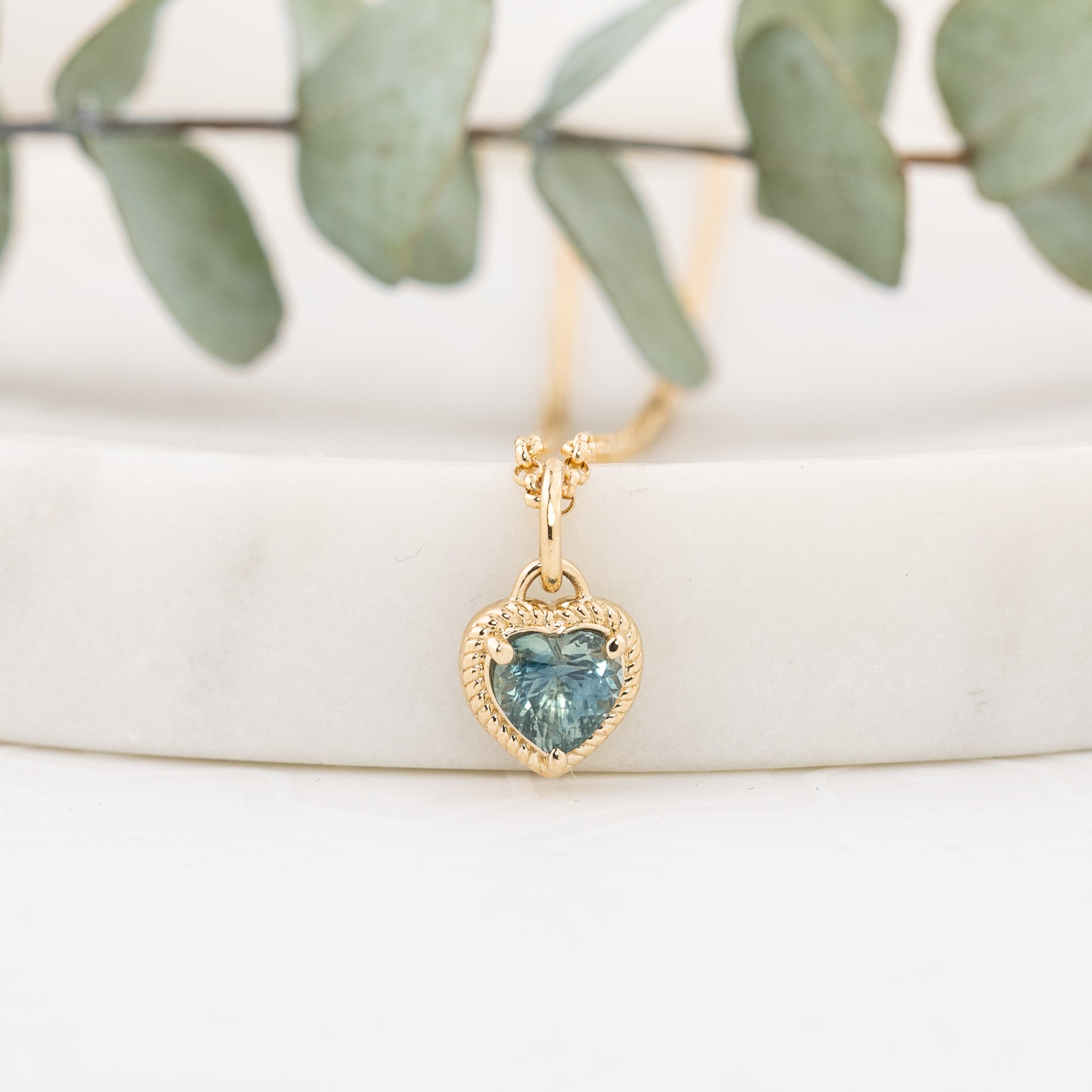 A gold heart-shaped pendant necklace with a blue heart-shaped gemstone, displayed on a white marble block with green eucalyptus leaves in the background.