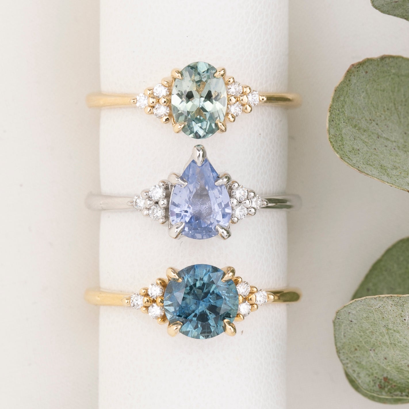 Three gold rings with different colored gemstones on a white surface with green leaves.