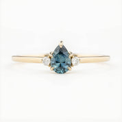 A pear-cut teal Montana sapphire ring with accent diamonds set in a 14k yellow gold band, displayed against a white background.