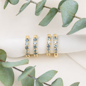 Three gold eternity rings with light blue gemstones are displayed on a white ribbon, framed by green eucalyptus leaves on a light beige background.