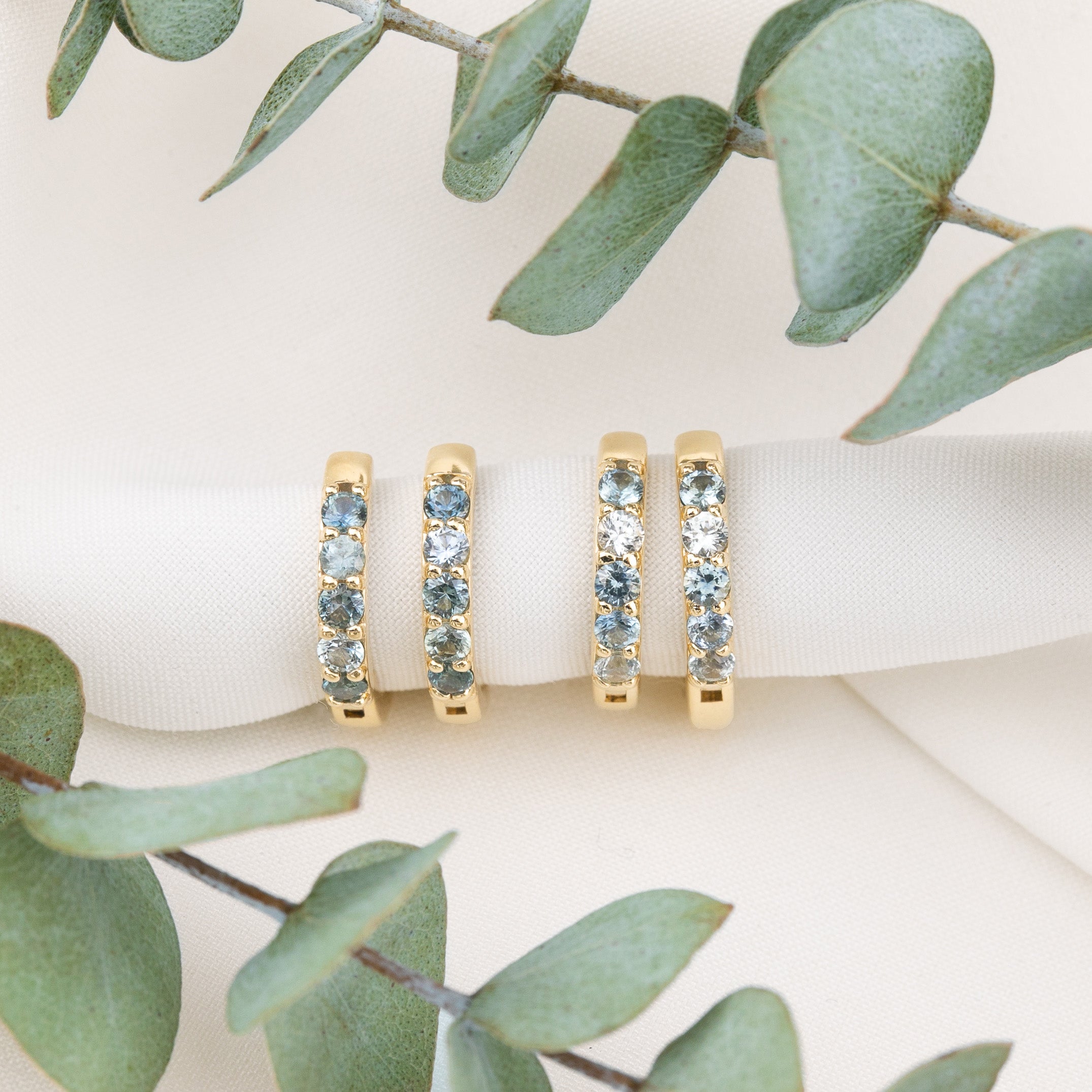 Three gold eternity rings with light blue gemstones are displayed on a white ribbon, framed by green eucalyptus leaves on a light beige background.