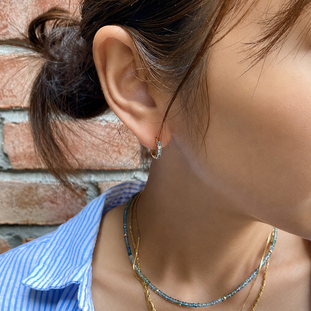 A close-up lifestyle shot of a woman wearing a small silver crescent moon stud earring with clear stones, styled with layered gold and blue beaded necklaces against a brick wall background.