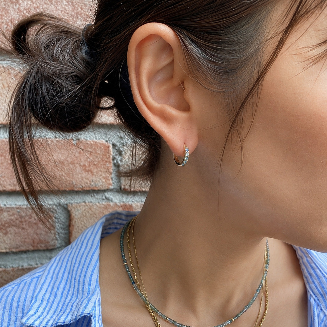 A close-up lifestyle shot of a woman's ear wearing a small silver crescent moon stud earring, with layered gold and blue beaded necklaces and a blue-and-white striped shirt against a brick wall background.