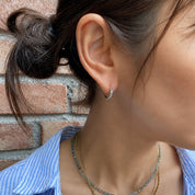 A close-up lifestyle shot of a woman's ear wearing a small silver crescent moon stud earring, with layered gold and blue beaded necklaces and a blue-and-white striped shirt against a brick wall background.