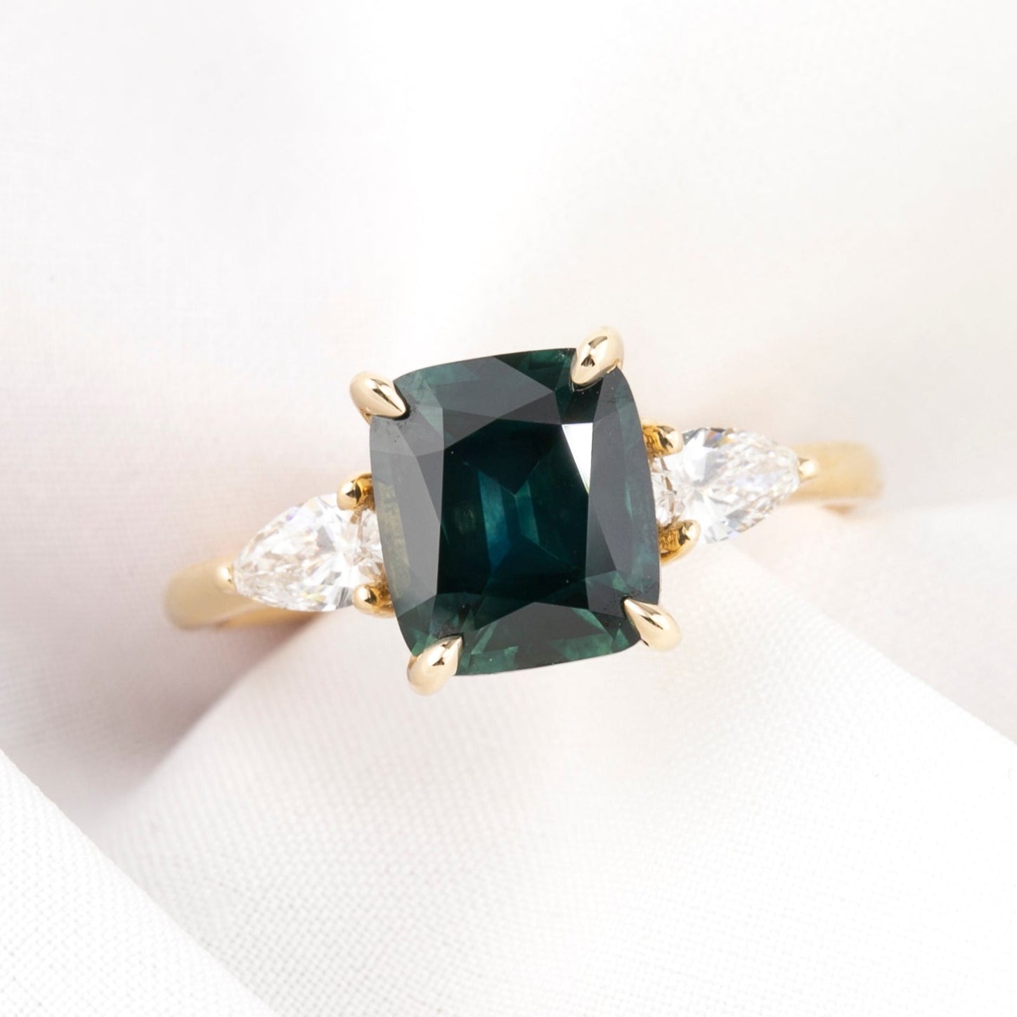 A cushion cut Montana Sapphire ring with deep blue green color, accompanied by natural accent diamonds, set in a 14k yellow gold band, displayed on a white background.