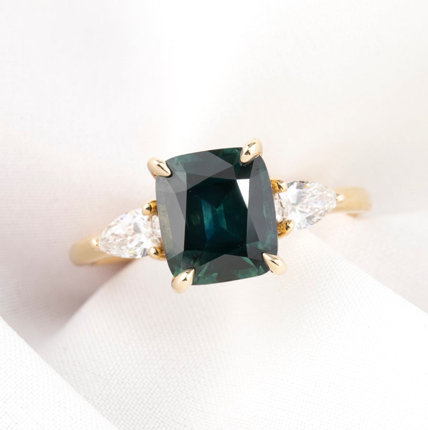 A cushion cut Montana Sapphire ring with deep blue green color, accompanied by natural accent diamonds, set in a 14k yellow gold band, displayed on a white background.