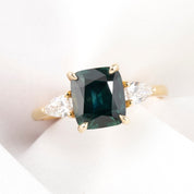 A cushion cut Montana Sapphire ring with deep blue green color, accompanied by natural accent diamonds, set in a 14k yellow gold band, displayed on a white background.
