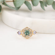 14k hexagonal gold ring with a central green round green Montana gemstone, surrounded by baguette and round accent diamonds and flanked by two pear-cut light purple gemstones. 