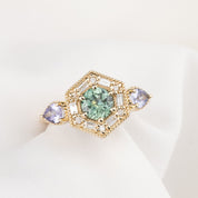 14k hexagonal gold ring with a central green round green Montana gemstone, surrounded by baguette and round accent diamonds and flanked by two pear-cut light purple gemstones. 