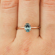 Close-up of a teal blue 1.00ct Montana sapphire set in a 14k rose gold ring on a finger.