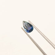1.06ct pear shaped dark blue Montana sapphire with deep royal navy color held by tweezers on white background