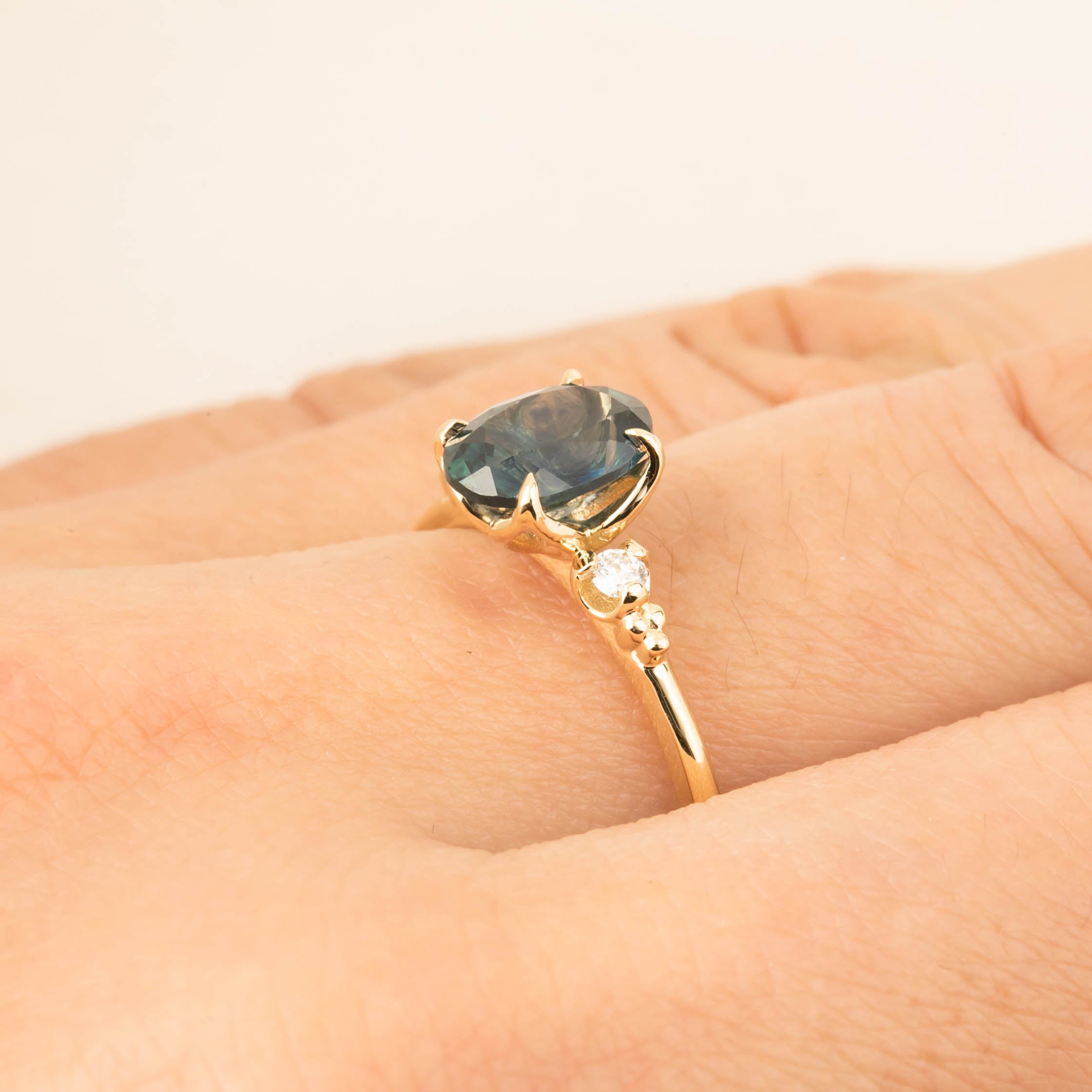 Close-up of a 14k gold ring with a 1.56ct teal blue Montana sapphire and small white accent diamonds on finger