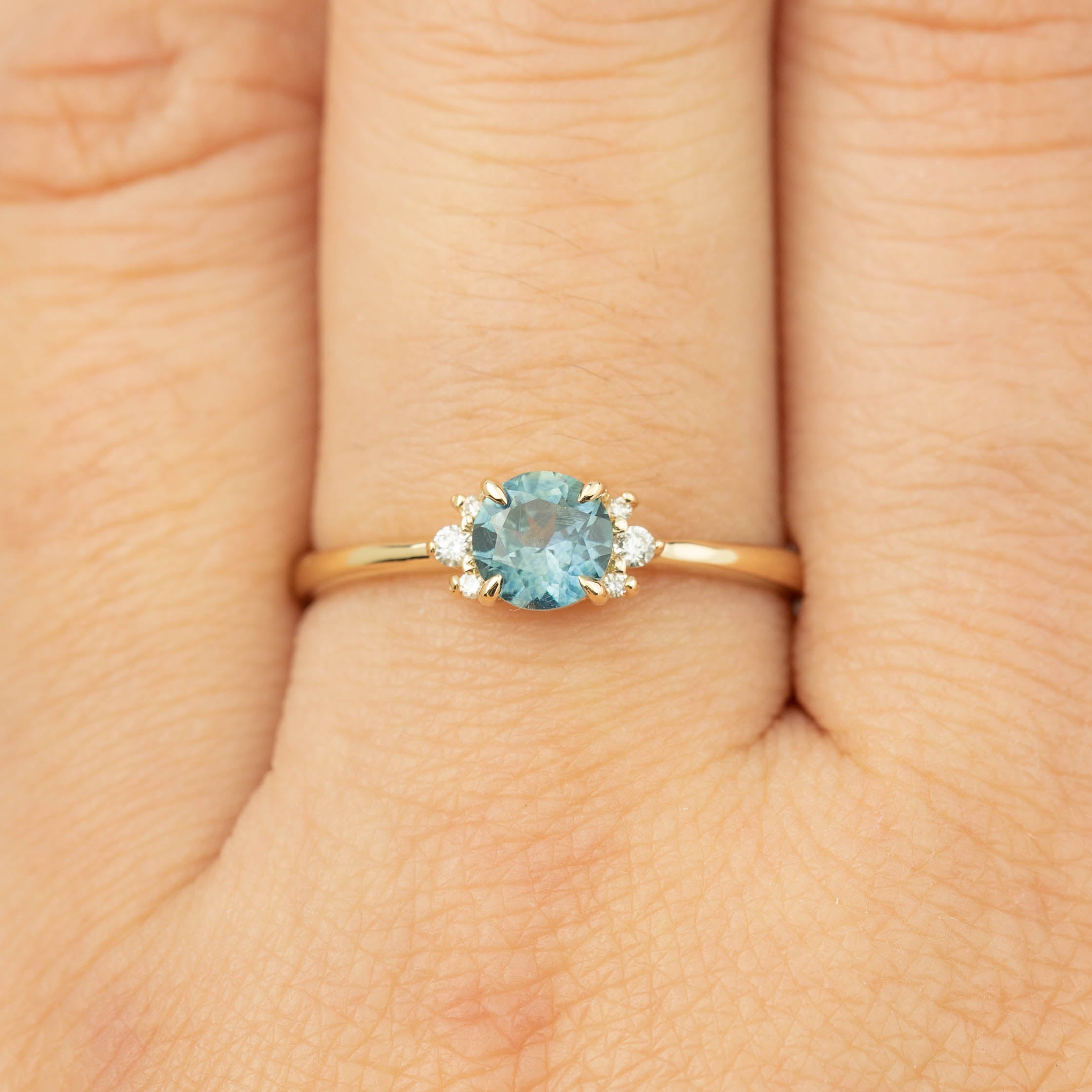 Close-up of a 14k gold ring with a 0.66ct teal Montana sapphire and small diamonds on a finger.