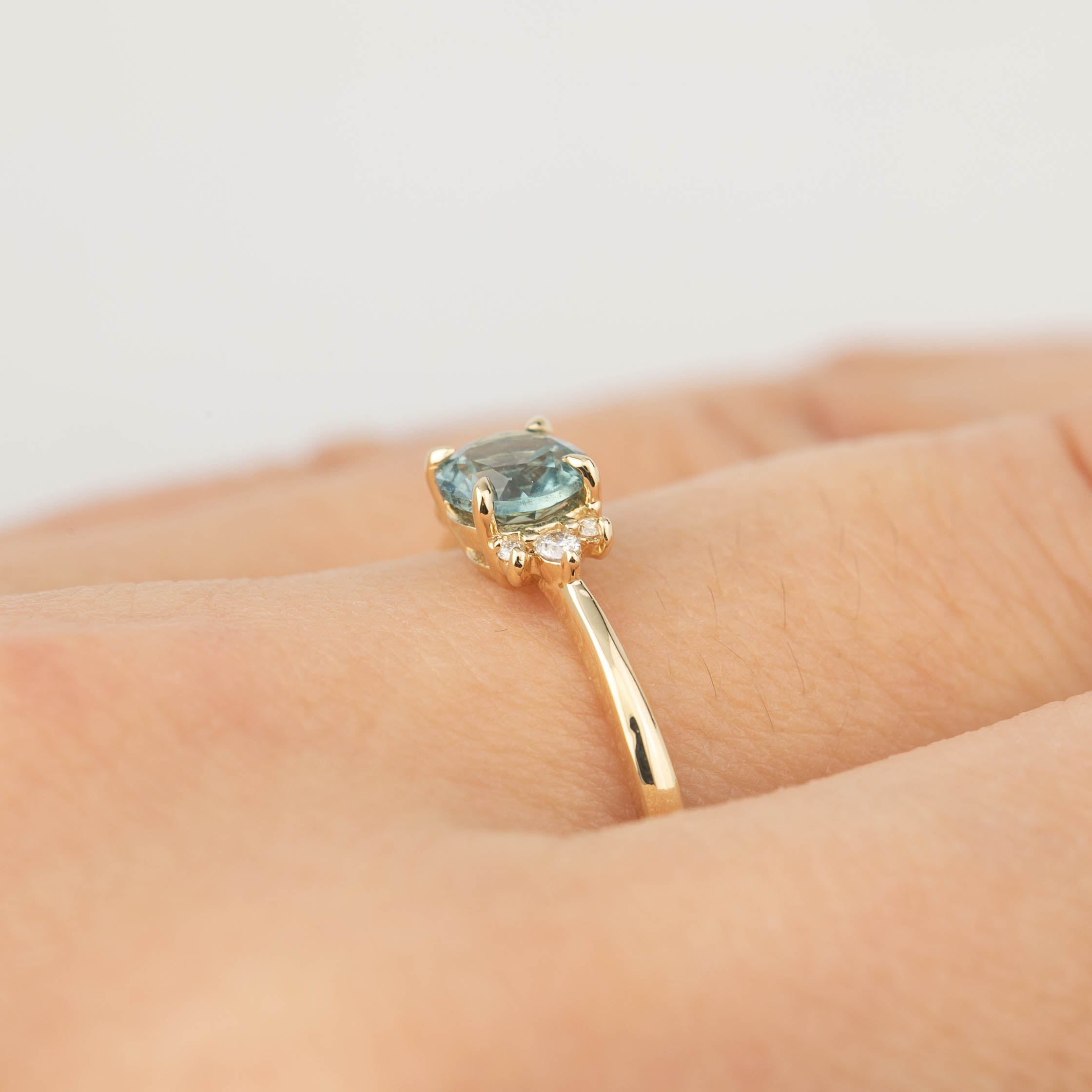 Close-up of a 14k gold ring with a 0.66ct teal Montana sapphire and small diamonds on a finger.