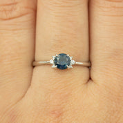 Close-up of a 0.65ct blue Montana sapphire ring in 14k white gold worn on a finger, featuring small diamonds.