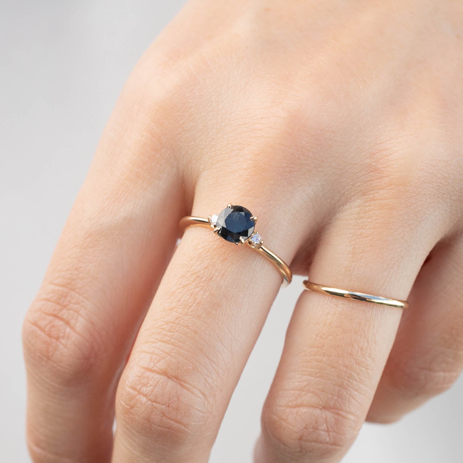 Hand wearing a 14k yellow gold ring with a 1ct dark peacock sapphire and small white diamond accent ring