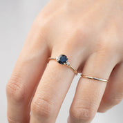Hand wearing a 14k yellow gold ring with a 1ct dark peacock sapphire and small white diamond accent ring