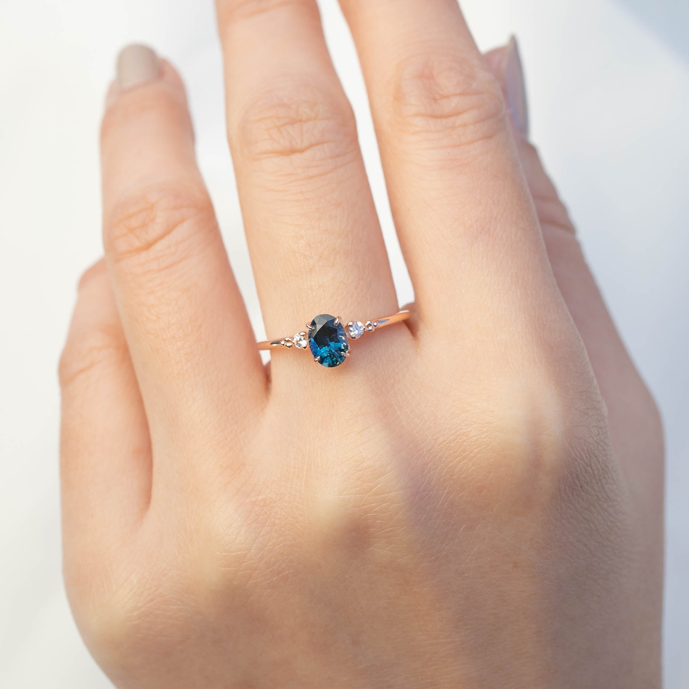 Hand wearing Estel Ring with 0.83ct teal blue sapphire and white accent diamonds in 14k rose gold band