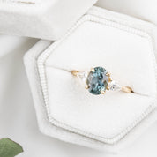 An oval-cut color-changing Montana sapphire ring with a yellow gold band and accent diamonds, displayed in a white hexagonal box with a green gemstone below.