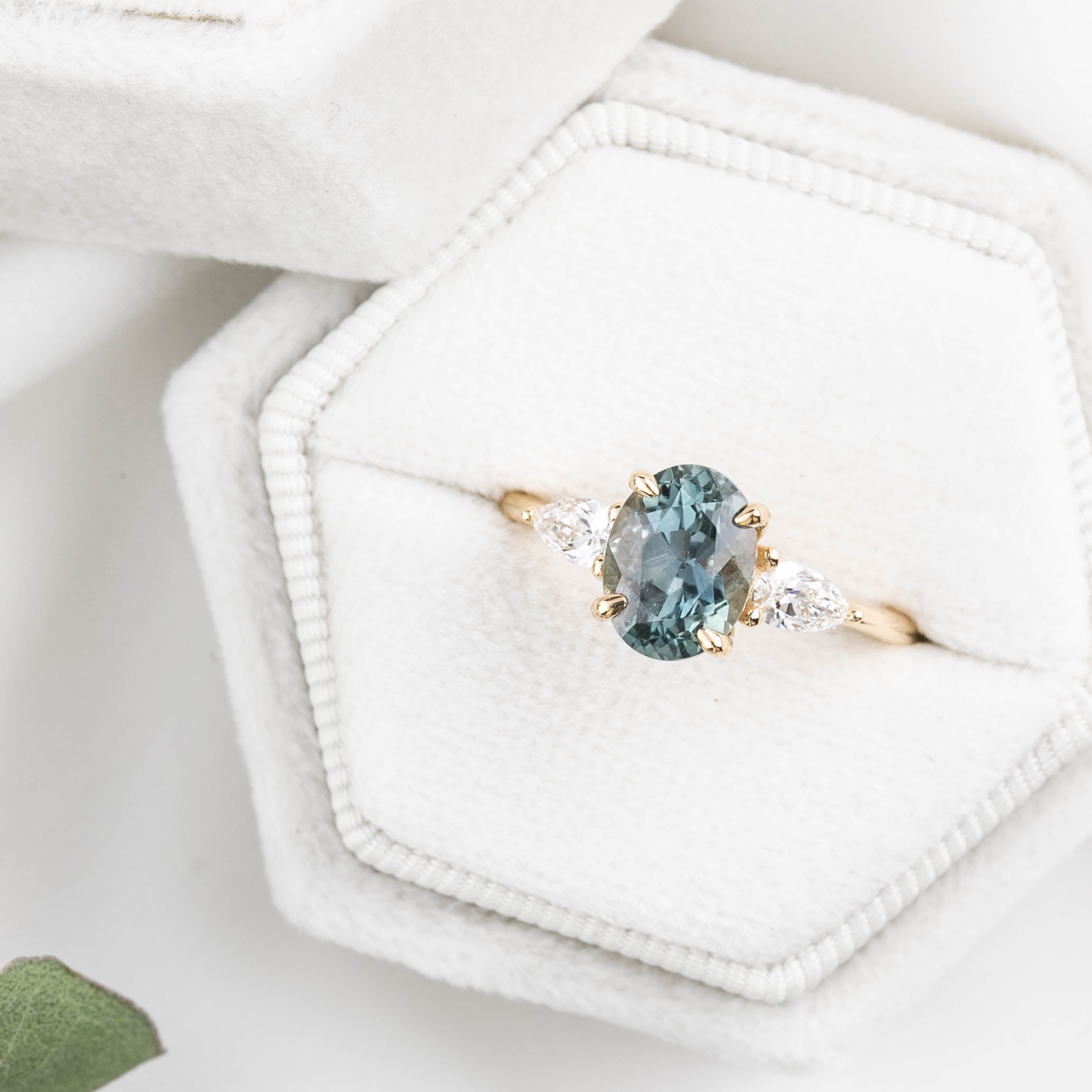An oval-cut color-changing Montana sapphire ring with a yellow gold band and accent diamonds, displayed in a white hexagonal box with a green gemstone below.