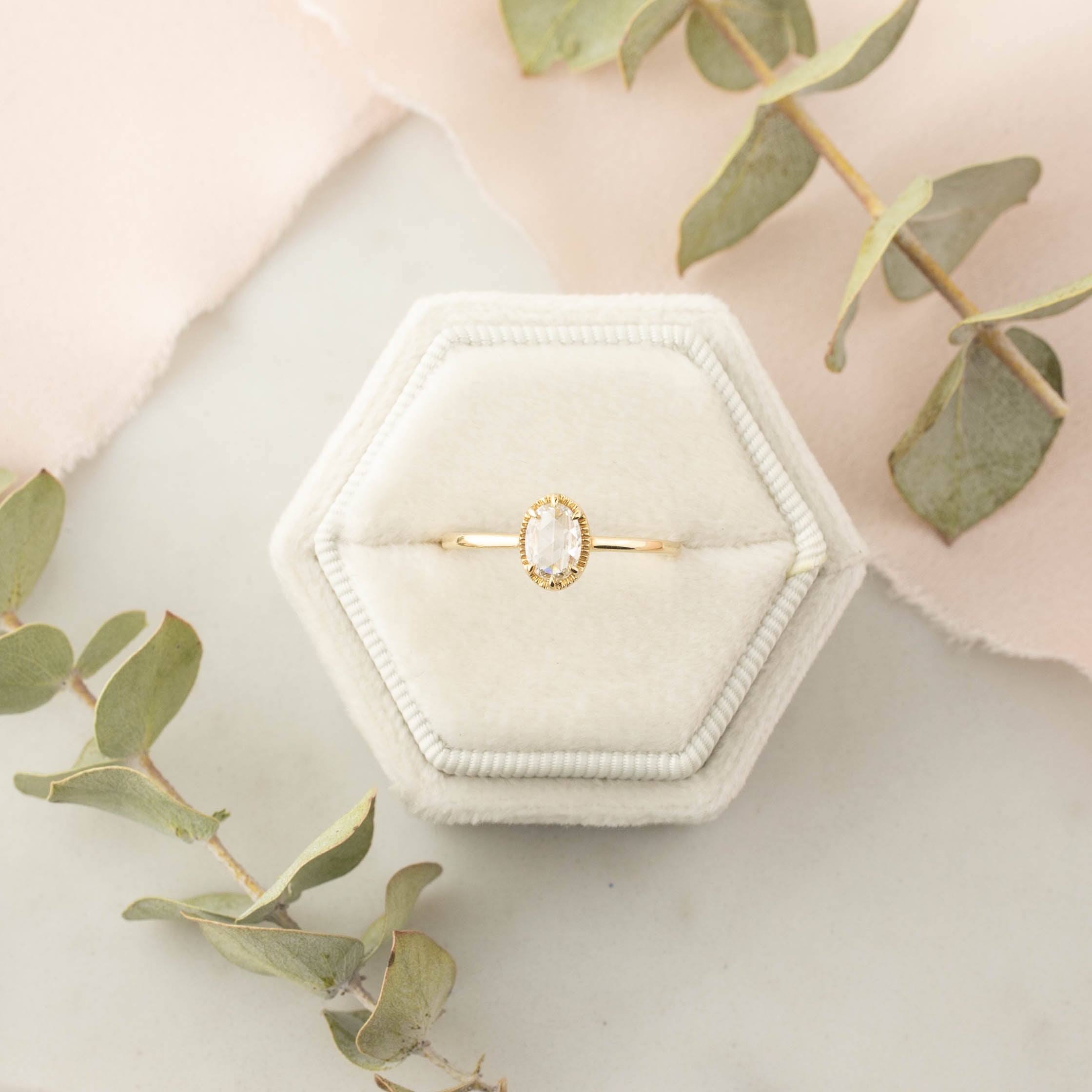 Oval rose cut diamond ring with gold tapered band in a hexagonal velvet box surrounded by green leaves