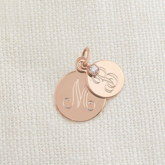 Two rose gold engraved circle pendants with a small gemstone on a textured fabric background