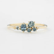 Three cluster rings with blue-green sapphires set in low profile prong settings, displayed on a white stand. The rings are crafted in recycled 14k solid gold.