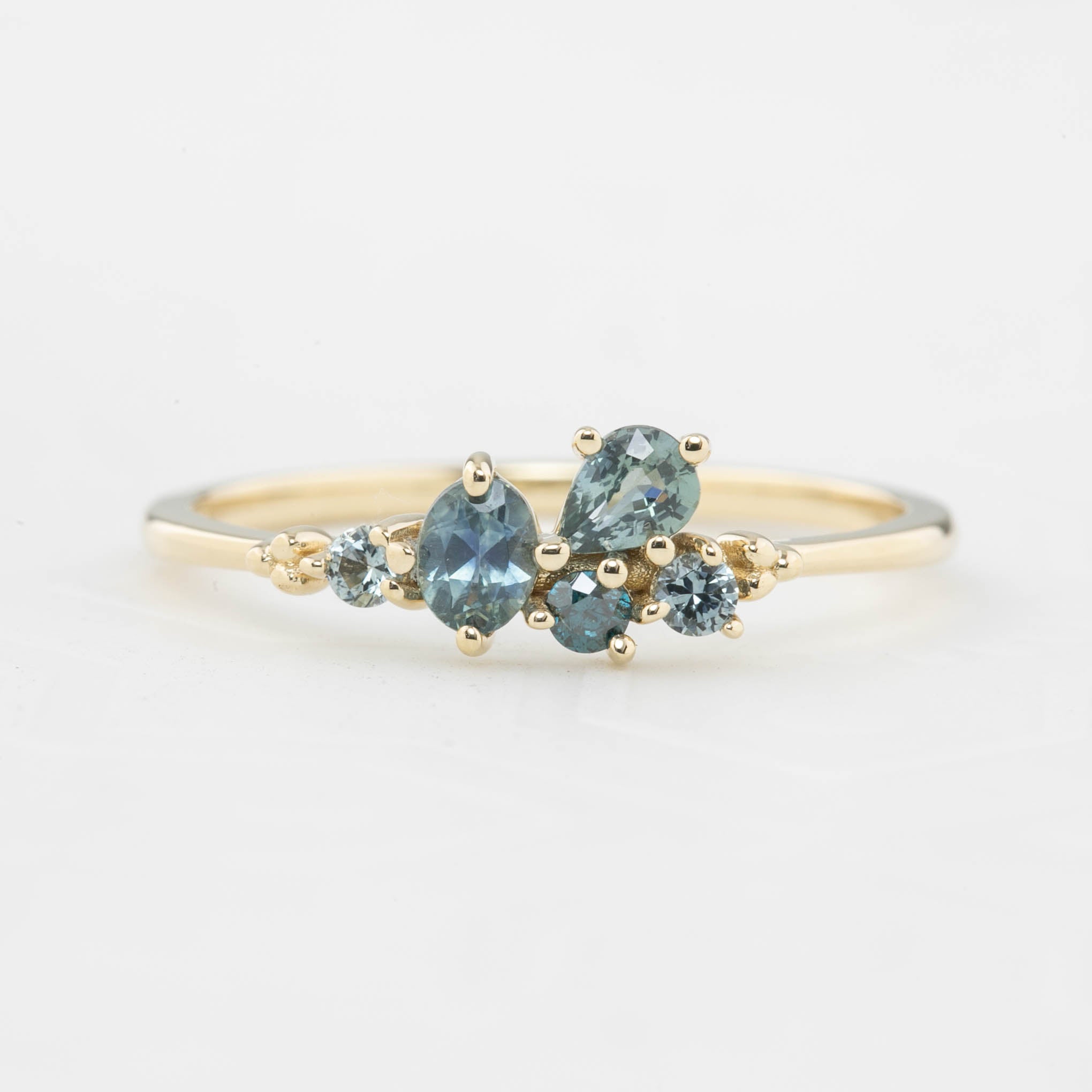 Three cluster rings with blue-green sapphires set in low profile prong settings, displayed on a white stand. The rings are crafted in recycled 14k solid gold.